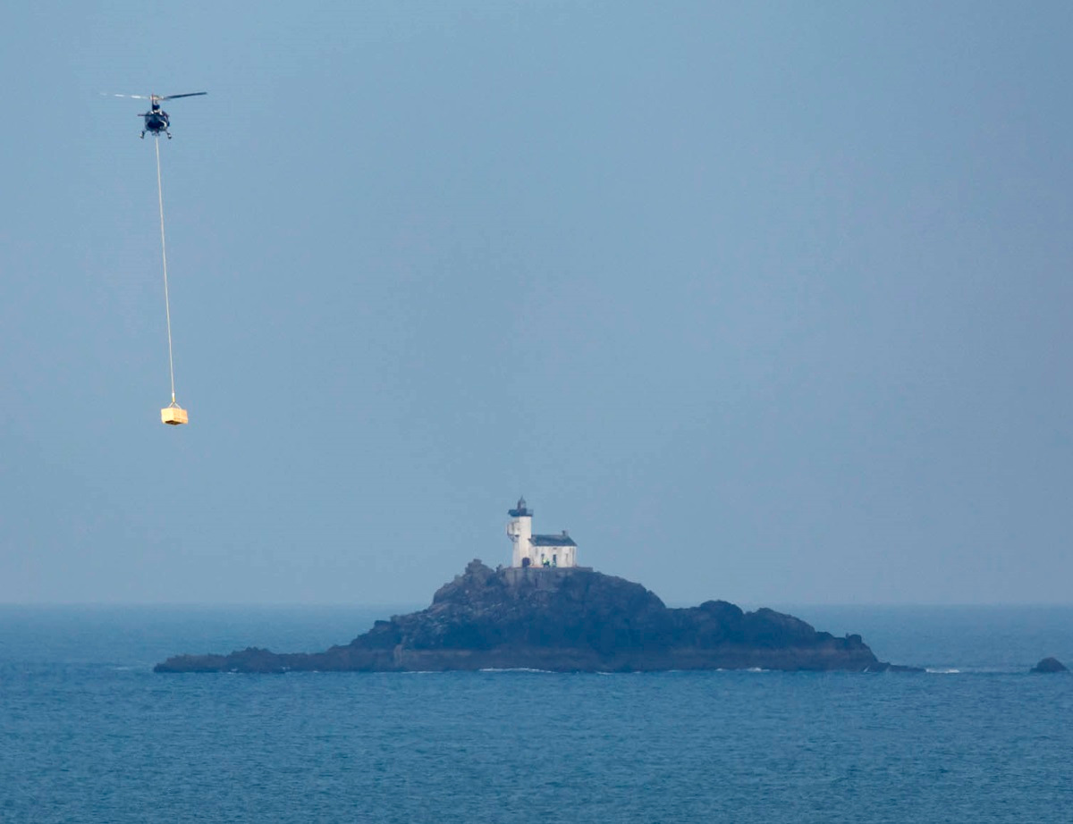Le phare de Tévénnec au loin, un hélicoptère apporte du matériel le long d'une corde.