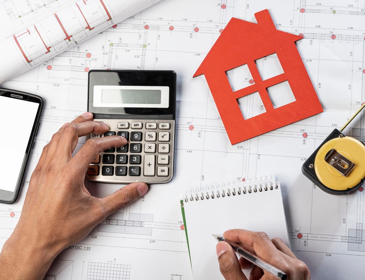 mains avec une calculatrice près d'un plan de maison